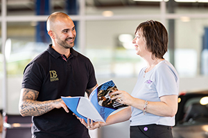 A Capricorn Risk Account Manager smiling while discussing a brochure with a Member inside an automotive showroom, with a car and office setting in the background.