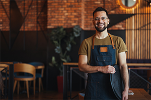 A cheerful small business owner wearing an apron, standing confidently in a stylish café with wooden and industrial décor.
