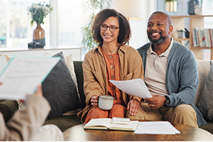 A smiling couple sitting on a couch, holding documents and a coffee mug, engaged in a conversation with a professional in a well-lit home setting.