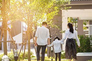 A family walking hand in hand towards their home, surrounded by greenery, with warm sunlight filtering through the trees.