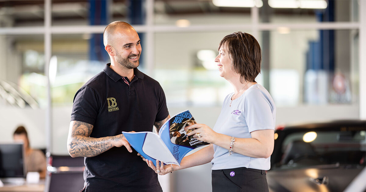 A Capricorn Risk Account Manager smiling while discussing a brochure with a Member inside an automotive showroom, with a car and office setting in the background.