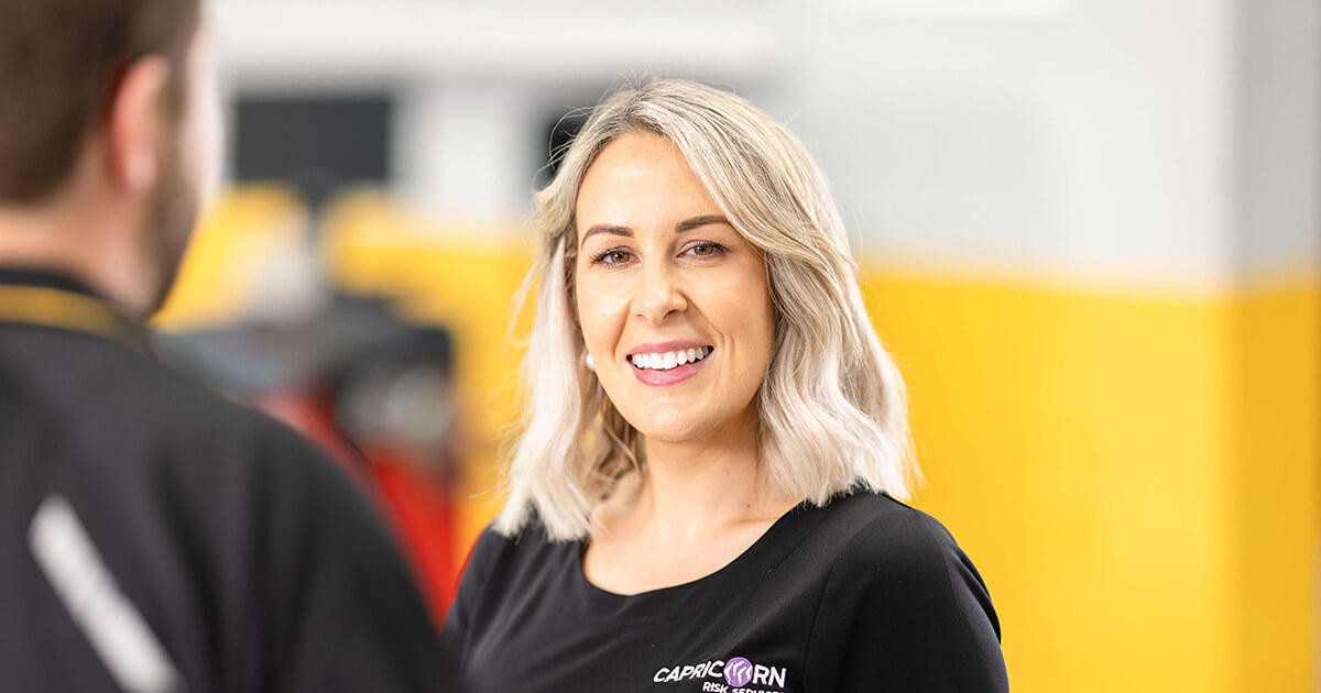 A Capricorn Risk Account Manager smiling while engaging in conversation with a Member in a workshop setting, with a blurred background featuring automotive equipment and yellow accents.