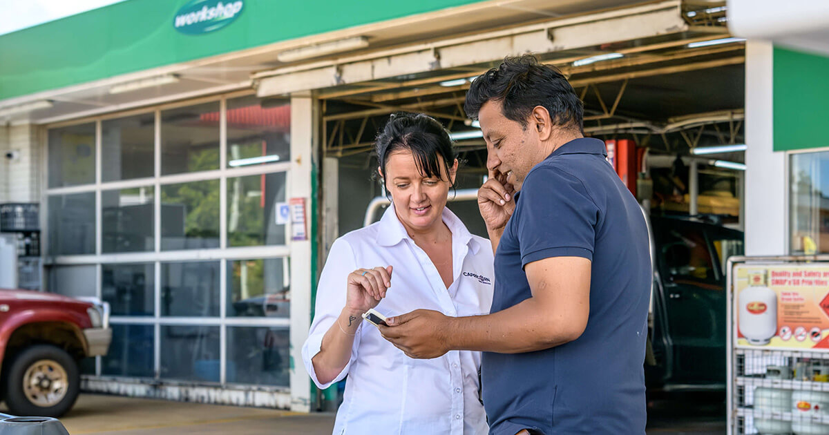 A Capricorn representative in a white shirt assisting a Member outside a workshop, both looking at a mobile device, with a green and white storefront in the background.